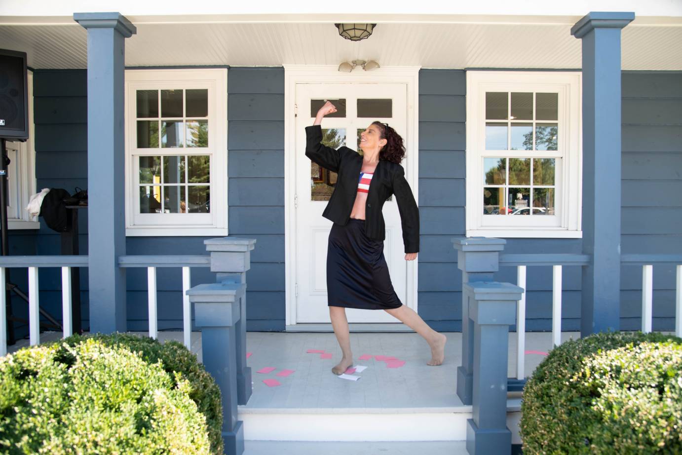 on the porch of a slate blue home with white trimmings a bare foot woman wearing a skirt suit over an American flag designed tube top, strikes a pose showing her strength, scattered around her feet are various index cards in pink and white